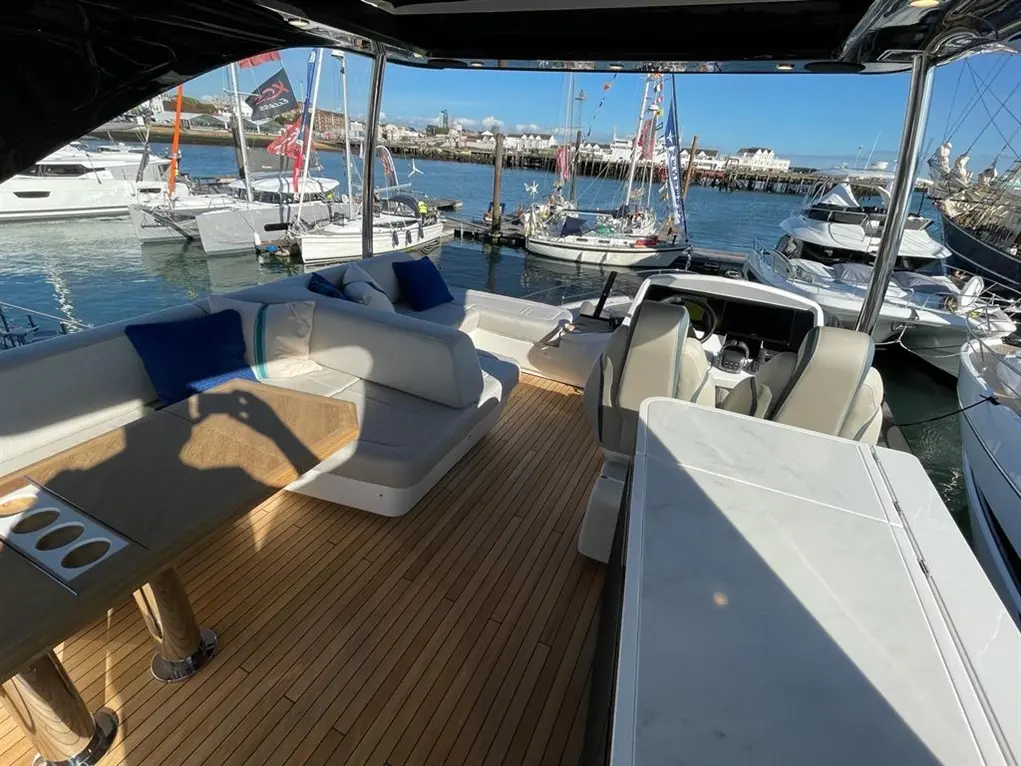 Luxury boat interior with wooden decking, seating area, and helm, featuring nearby yachts in a marina with clear skies.